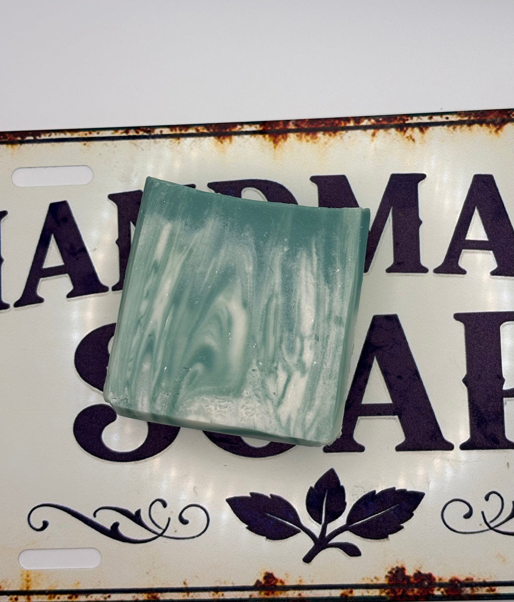 A rectangular bar of handmade soap with a swirled mint green and white pattern, resting on a vintage-style white metal sign with dark purple lettering.