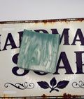 A rectangular bar of handmade soap with a swirled mint green and white pattern, resting on a vintage-style white metal sign with dark purple lettering.
