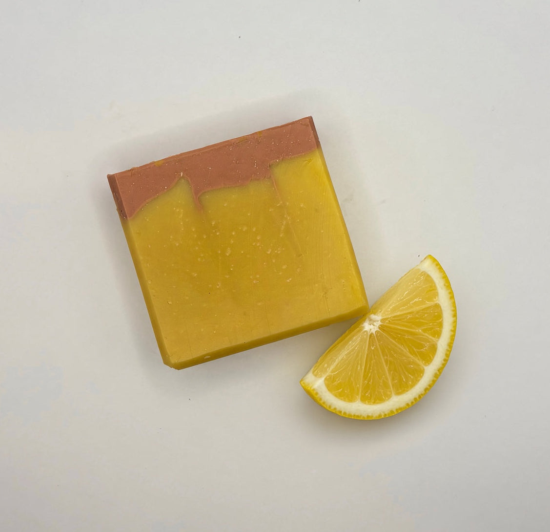 A rectangular bar of yellow soap with a copper-brown top layer, photographed on a plain light background next to a lemon slice.