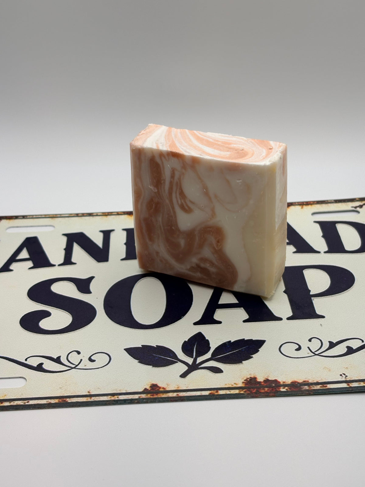 A rectangular bar of handmade soap with a marbled cream and peach swirl pattern, standing upright on a vintage-style metal sign that reads 'HANDMADE SOAP'.