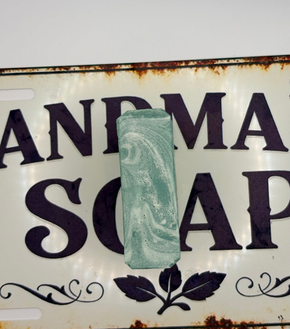 A rectangular bar of handmade soap with a light green and white marbled swirl pattern, displayed on a vintage-style metal sign that reads 'HANDMADE SOAP' with rusted edges.