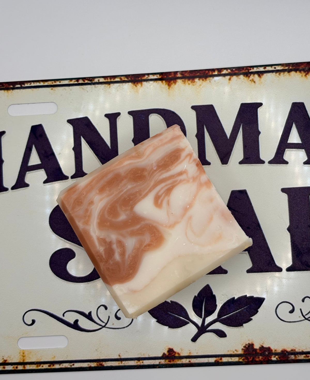 A rectangular bar of marbled soap in peach and cream tones, placed on a decorative vintage-style metal sign with the words 'HANDMADE SOAP'.
