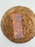 A rectangular handmade soap bar with a marbled swirl pattern of orange, purple, and white, displayed on a round woven jute placemat against a plain white background.