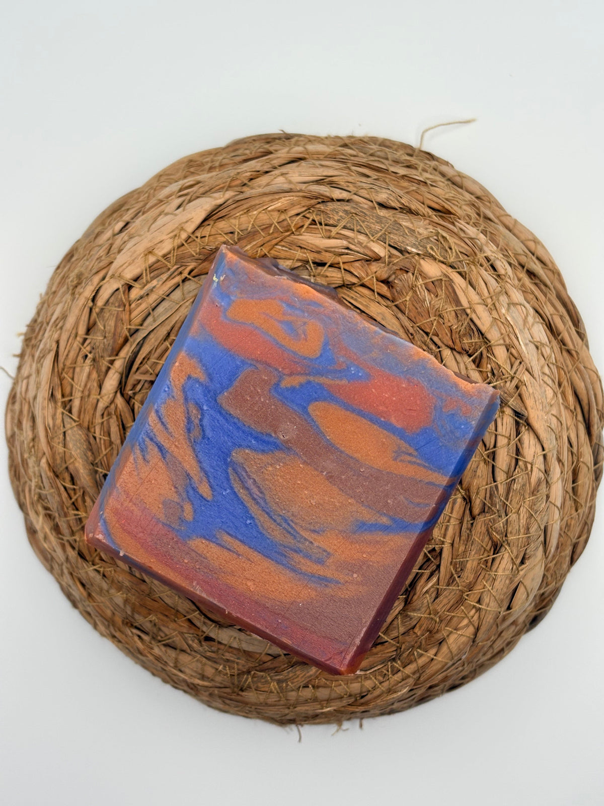 A square handmade soap bar with a marbled pattern of blue, rust orange, and deep red, displayed on a round woven straw placemat against a white background.