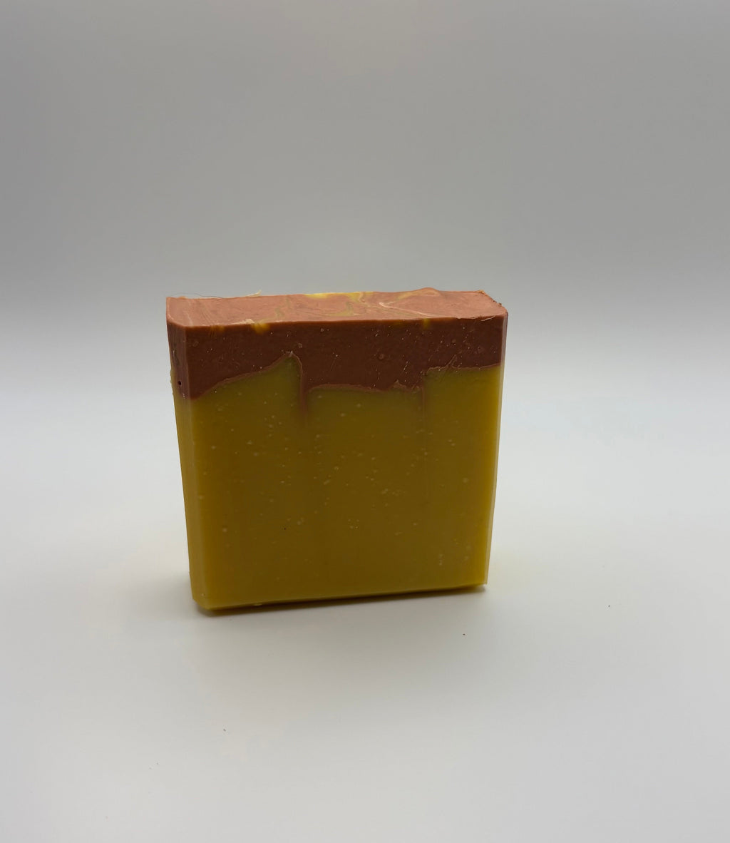 A rectangular bar of handmade soap with a two-tone design, featuring a golden yellow lower half and a reddish-brown top layer, set against a neutral gray studio background.