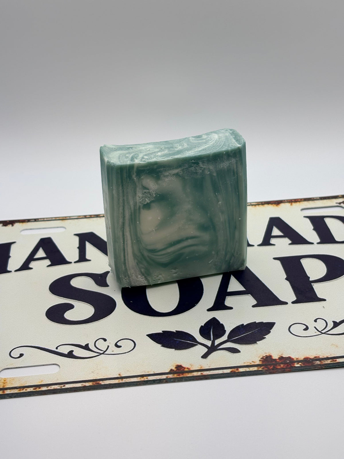A rectangular bar of handmade soap with a green and white marbled face design, standing upright on a decorative vintage-style metal sign that reads 'Handmade Soap'.
