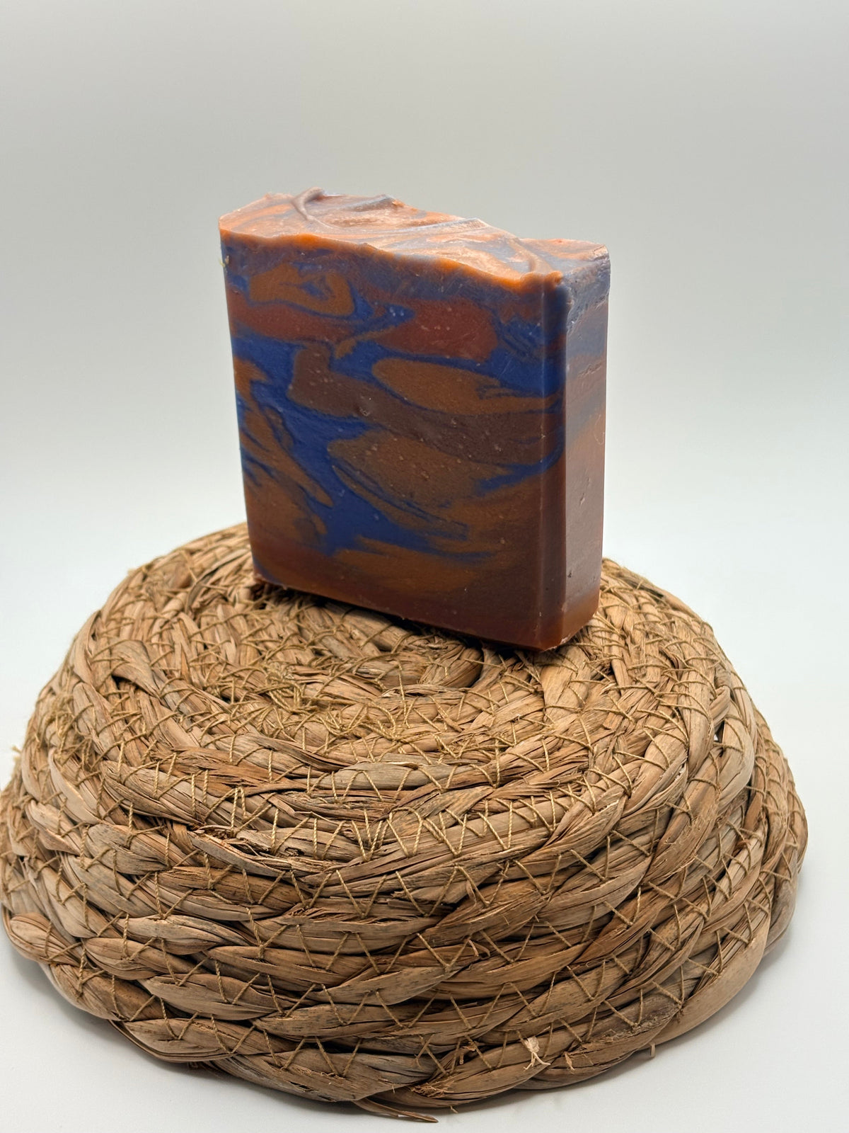 A rectangular bar of marbled soap in orange-brown and blue, displayed upright on a round woven straw holder against a plain light gray background.