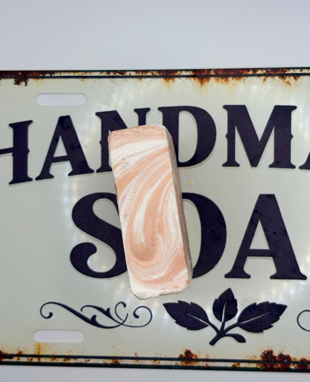 A rectangular bar of soap with a peach and white marble swirl pattern, displayed on a vintage-style metal license plate that reads 'HANDMADE SODA'.