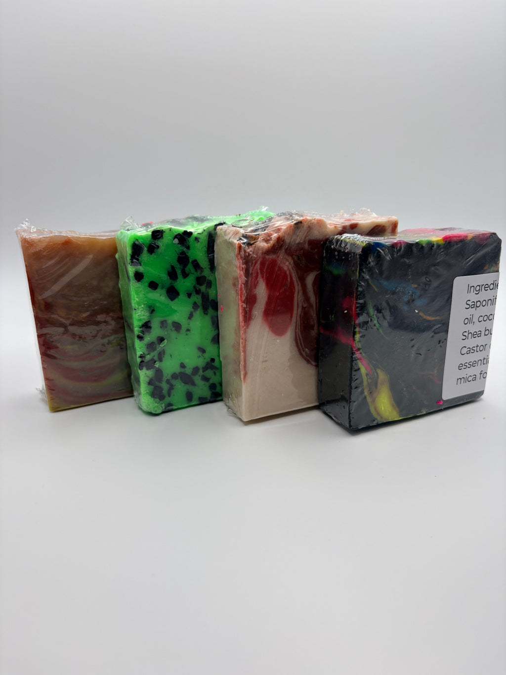 Four handmade soap bars wrapped in clear plastic, arranged in a row on a light gray background. The soaps have swirled patterns in brown, bright green with black speckles, beige with red swirls, and black with red, yellow, and blue swirls.