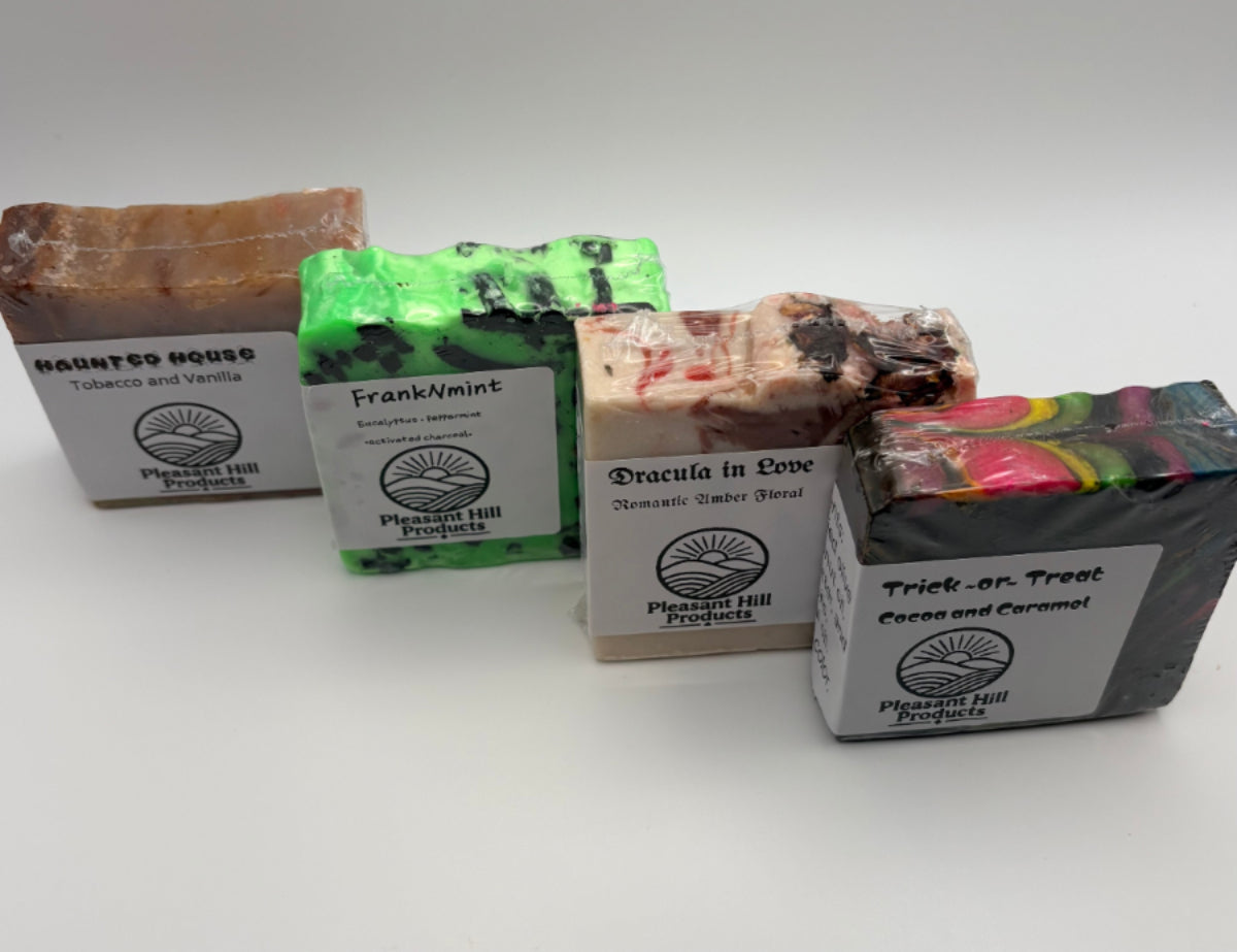 Four artisanal bars of soap from Pleasant Hill Products arranged on a white background. The soaps include Tobacco and Vanilla, Frankinmint, Dracula in Love, and Trick or Treat Cacao and Coffee.