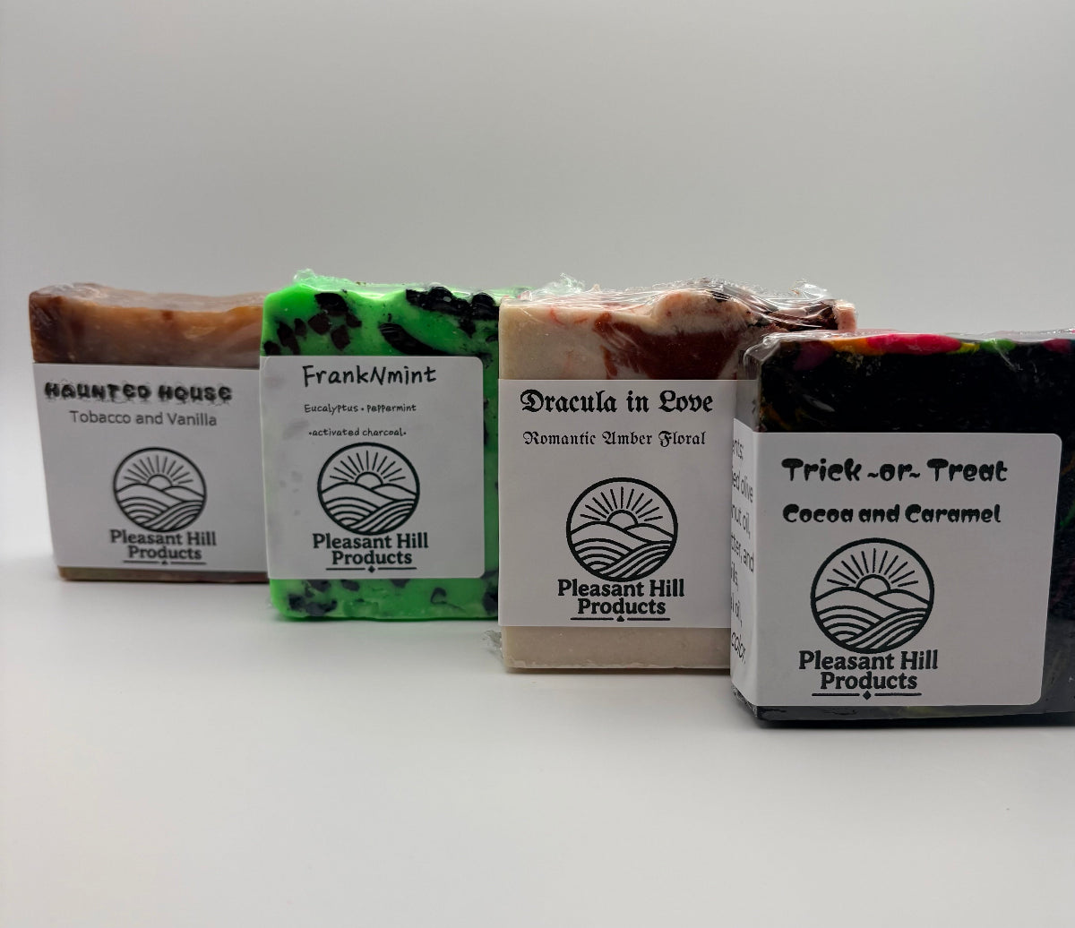 Four bars of Pleasant Hill Products artisan soap arranged in a row on a white background. The soaps have colorful marbled tops and white labels with black text, showing scents: Haunted House (Tobacco and Vanilla), Frank/A Mint, Dracula in Love (Necromantic Amber Floral), and Trick or Treat (Cocoa and Caramel).